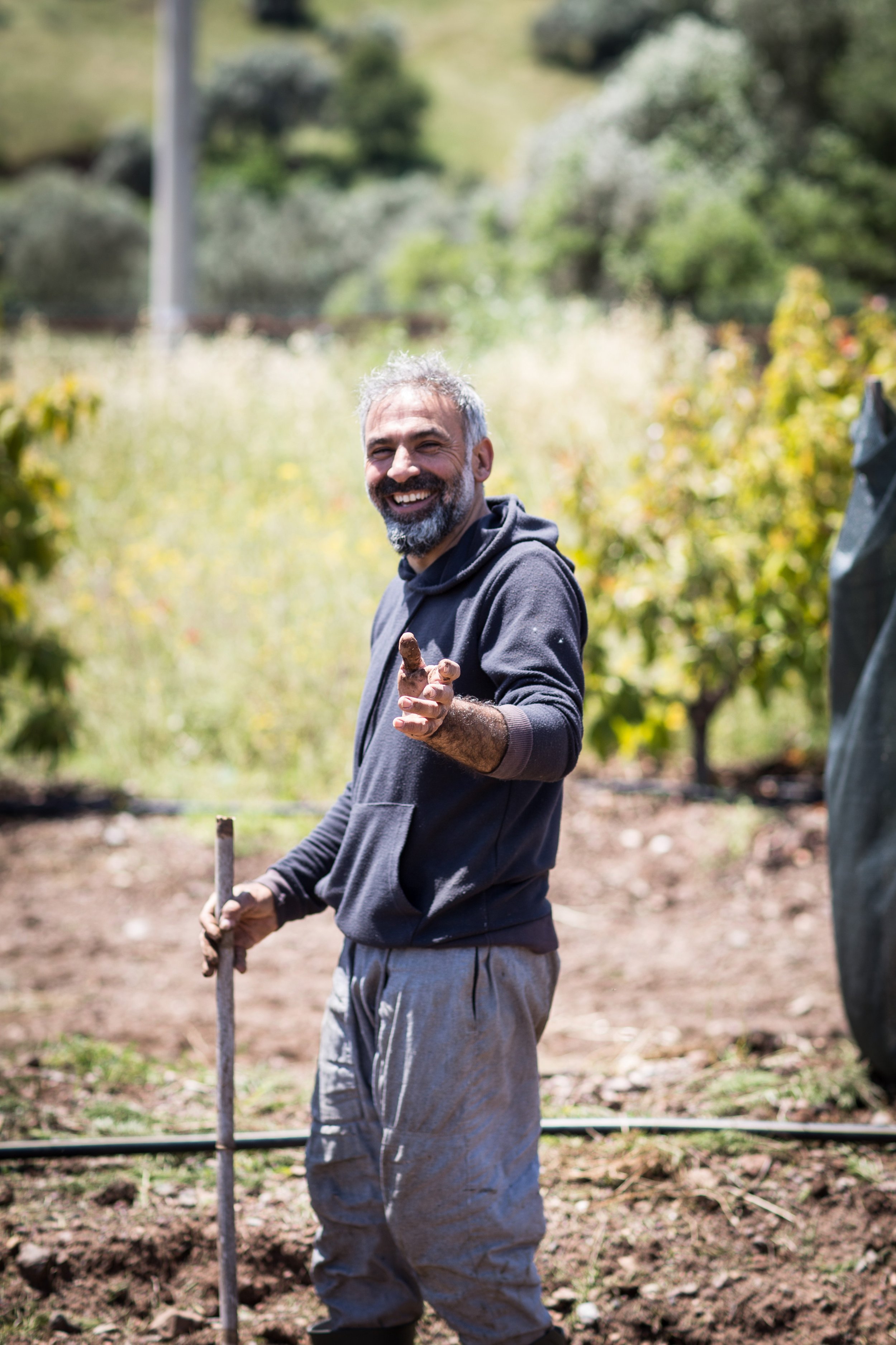 Ein lachender Mann mit grauem Bart und grauen Haaren hält einen Stock in der Hand und zeigt mit dem Finger auf die Kamera, während er in einem Garten steht.