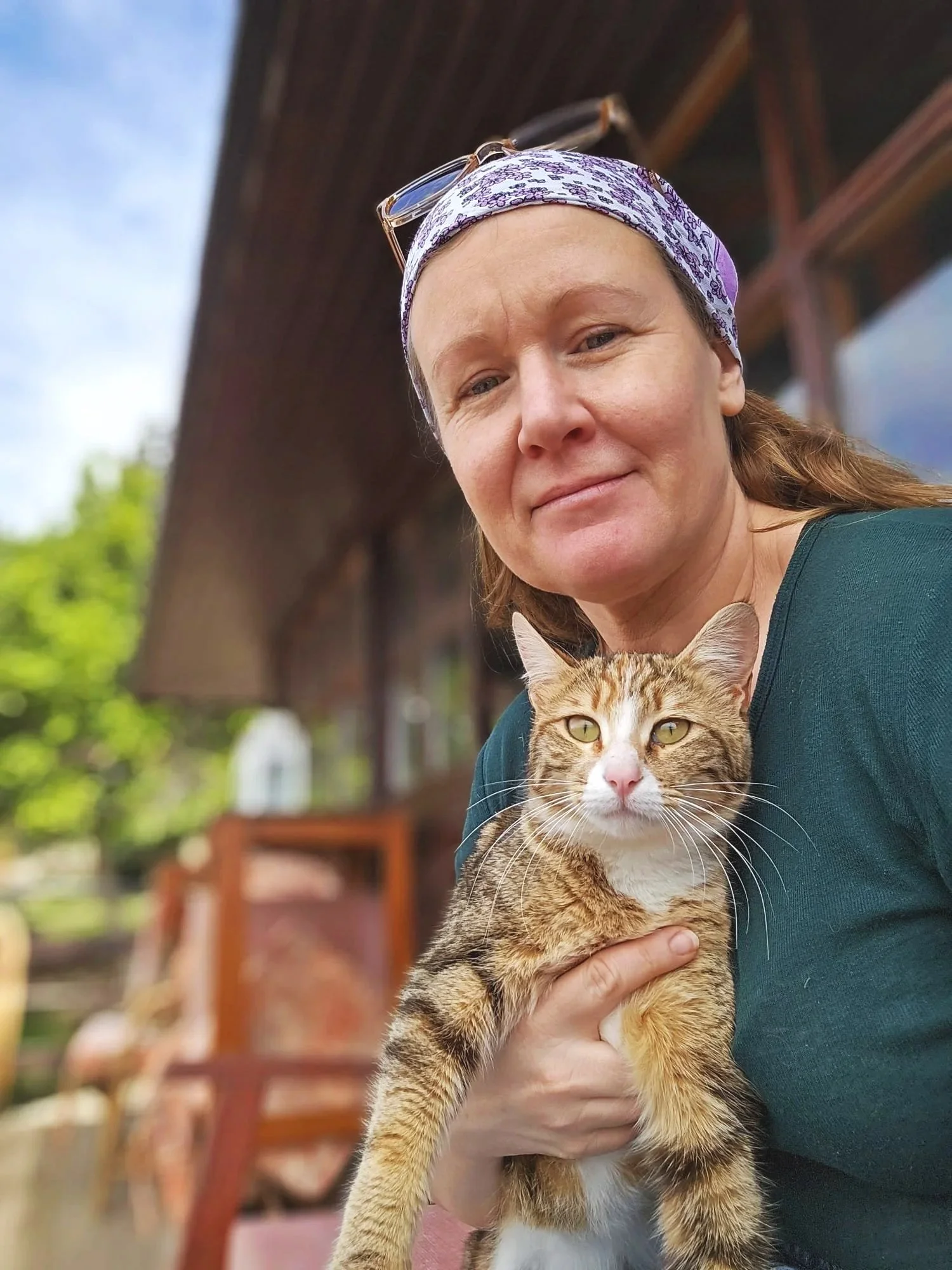 Eine Frau hält eine getigerte Katze im Freien. Die Frau trägt eine Brille auf dem Kopf und ein Tuch um den Kopf. Der Hintergrund zeigt einen Holzzaun und Bäume.