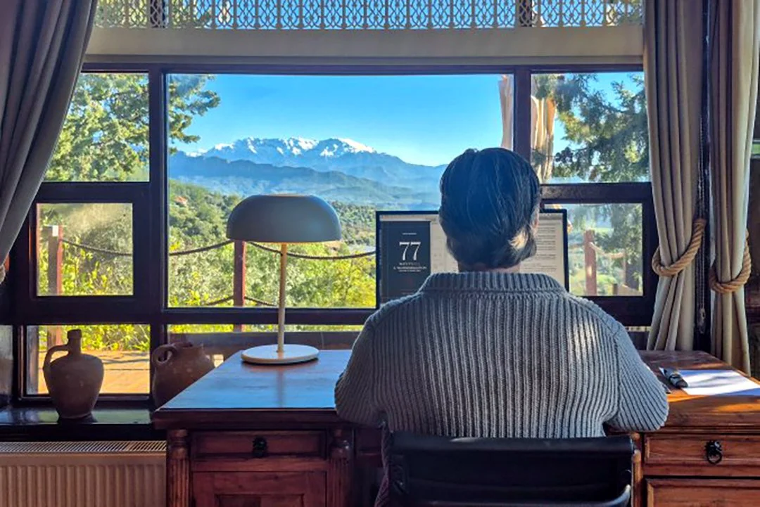 Person arbeitet an einem Schreibtisch vor einem großen Fenster mit Blick auf Berge und Natur, mit Keramikkannen und einer Lampe auf dem Tisch.