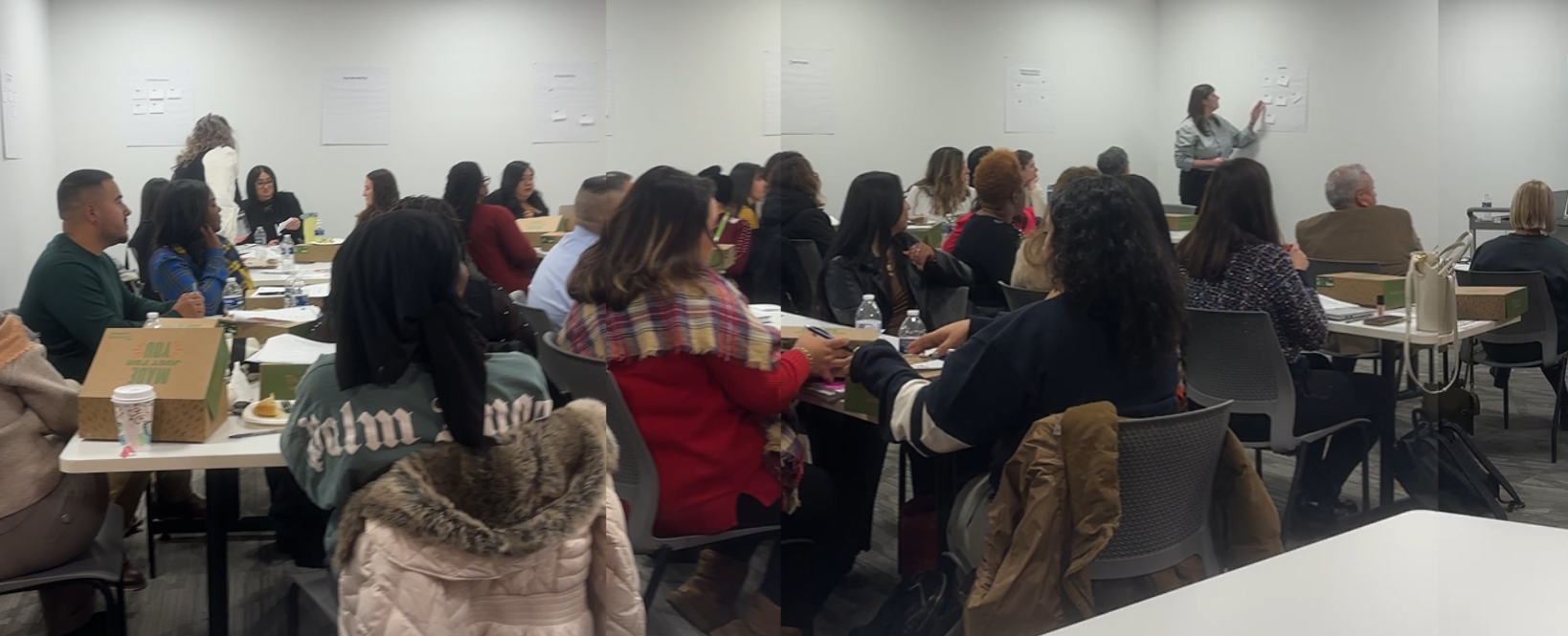 A group of diverse people attending a seminar or workshop in a conference room. They are seated at tables with notebooks, water bottles, and food, listening to a woman standing and presenting at the front of the room.