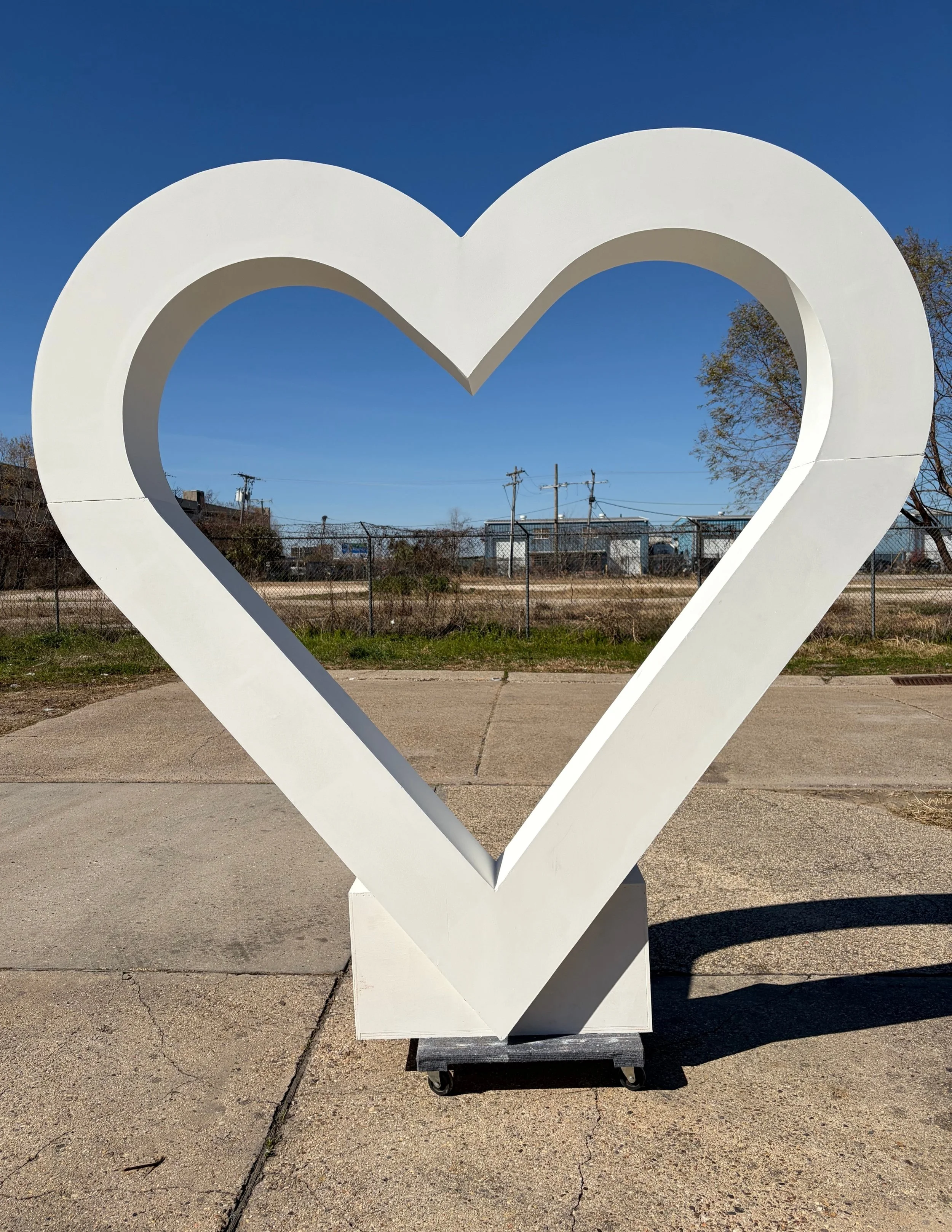 A large white heart-shaped sculpture outdoors on a sunny day, with trees, power lines, and a fence in the background.