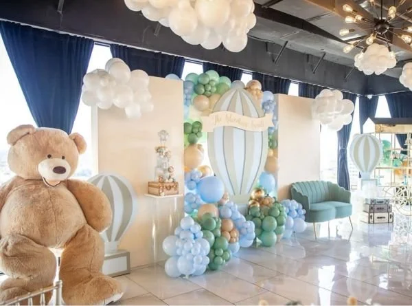 Baby shower celebration setup with pastel-colored balloons, a large teddy bear, a vintage stroller, and pastel blue and white decorations, including a hot air balloon motif.