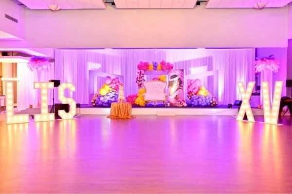Decorated event stage with large illuminated letters 'LIS' and 'XV', purple curtains, floral arrangements, and a central pink and white ornate chair.