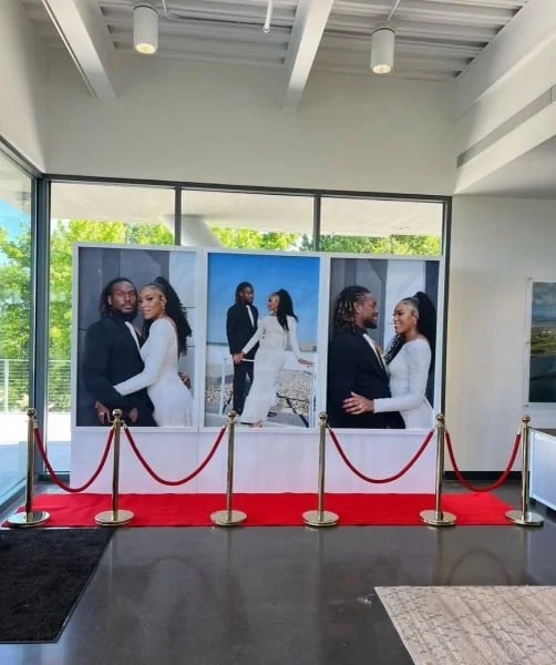 Display of large photographs of a couple in wedding attire, with a red carpet and stanchions, inside a modern building with large windows and a view of trees outside.