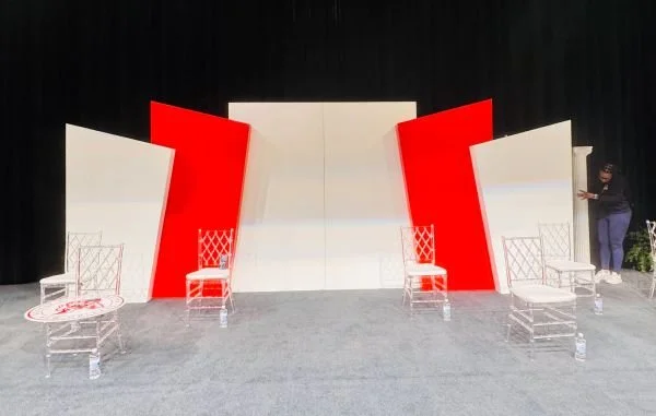 Stage setup with white and red geometric backdrops, four transparent chairs, and matching tables, with a person preparing the scene.