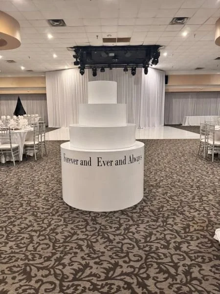 White wedding cake with three tiers on a round table in a large event hall, with draped white curtains and round tables with white chairs in the background.