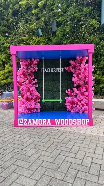Pink outdoor swing with a green seat, surrounded by pink flowering bushes, under a pink frame. Background features green trees, with text "TEACHER FEST" and "ZAMORA WOODSHOP" at the bottom.