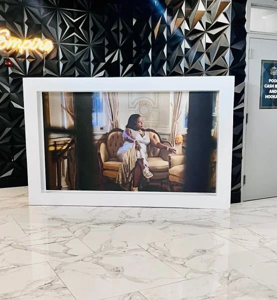 A large framed photograph of a woman sitting on a vintage-style sofa in a luxurious room, displayed on a marble floor in front of a black geometric wall.