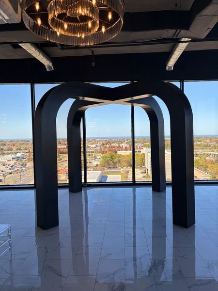 Modern black arch sculpture inside a high-rise building with large windows showing a cityscape and sky outside.