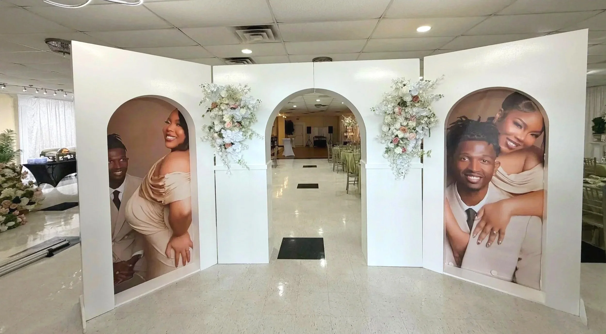 Wedding reception decor with a white floral arch featuring large portraits of a smiling couple, arranged in an indoor banquet hall with tables and chairs.