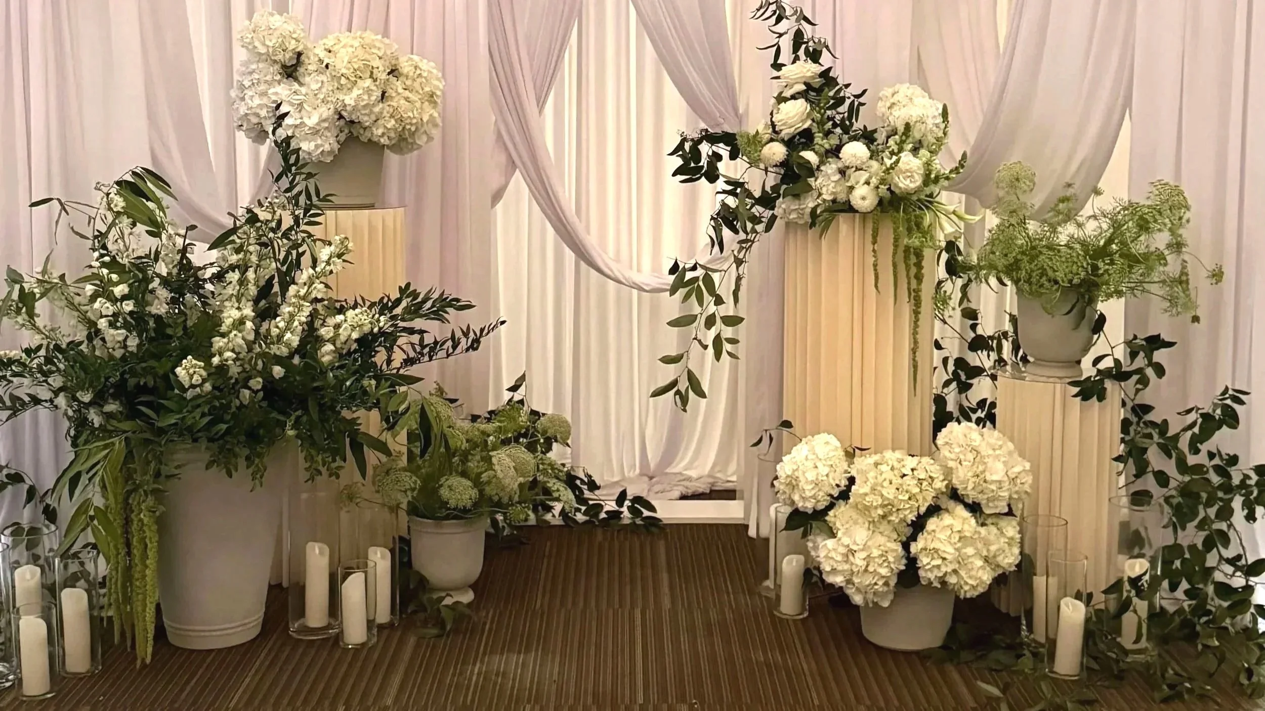 Elegant wedding altar decorated with white flowers, hydrangeas, roses, and greenery, with draped white curtains in the background and white candles along the floor.