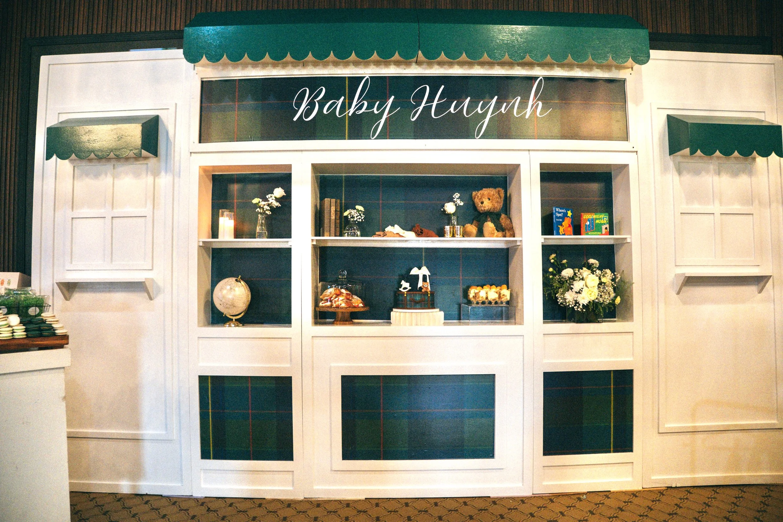 A white display booth with green scalloped awnings and a sign that reads 'Baby Haynh.' The booth contains decorative items including a teddy bear, small floral arrangements, books, a candle, and a globe, with a wooden wall background.