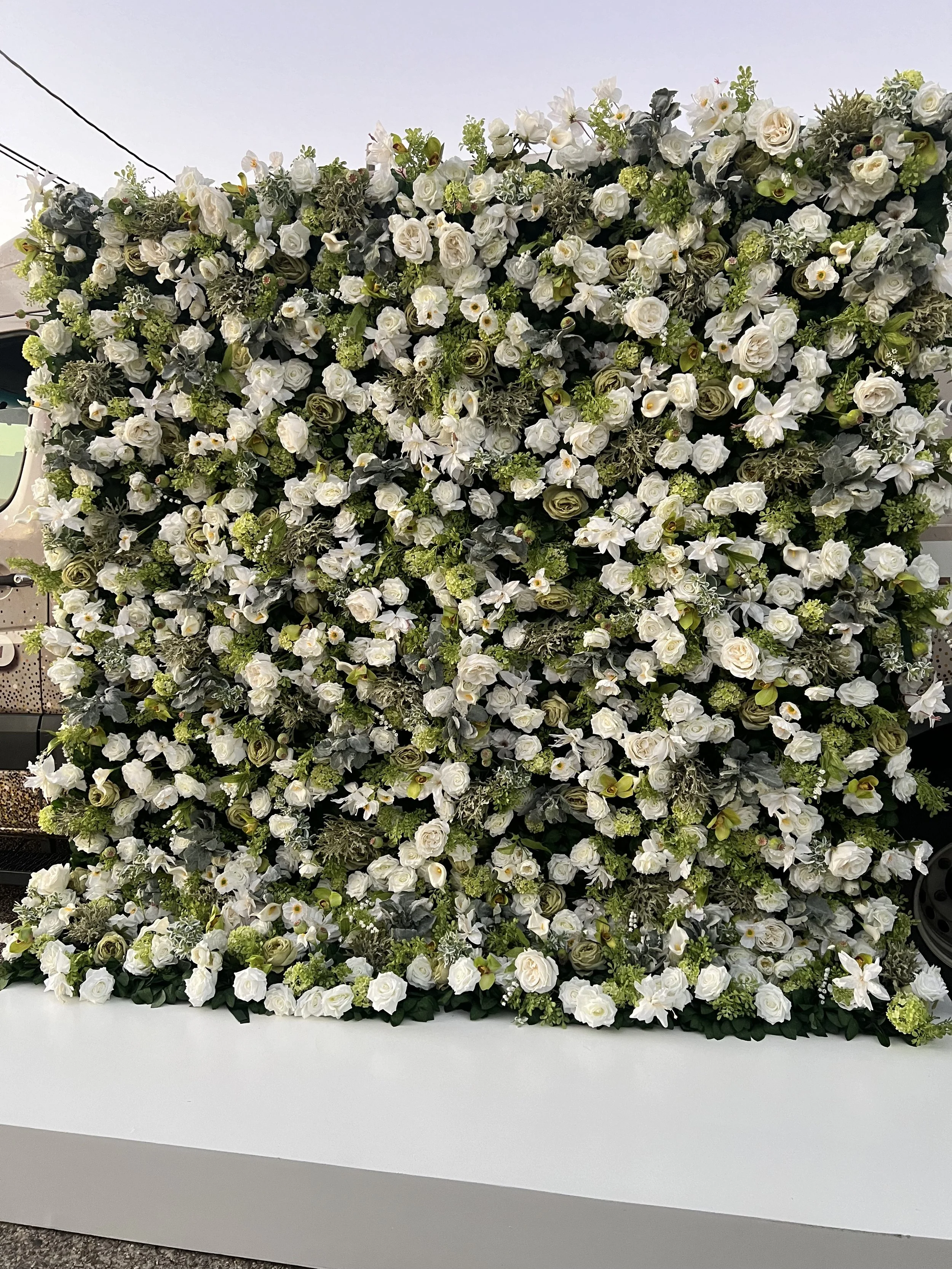A large rectangular arrangement of white roses, lilies, and various greenery, displayed on a white table.