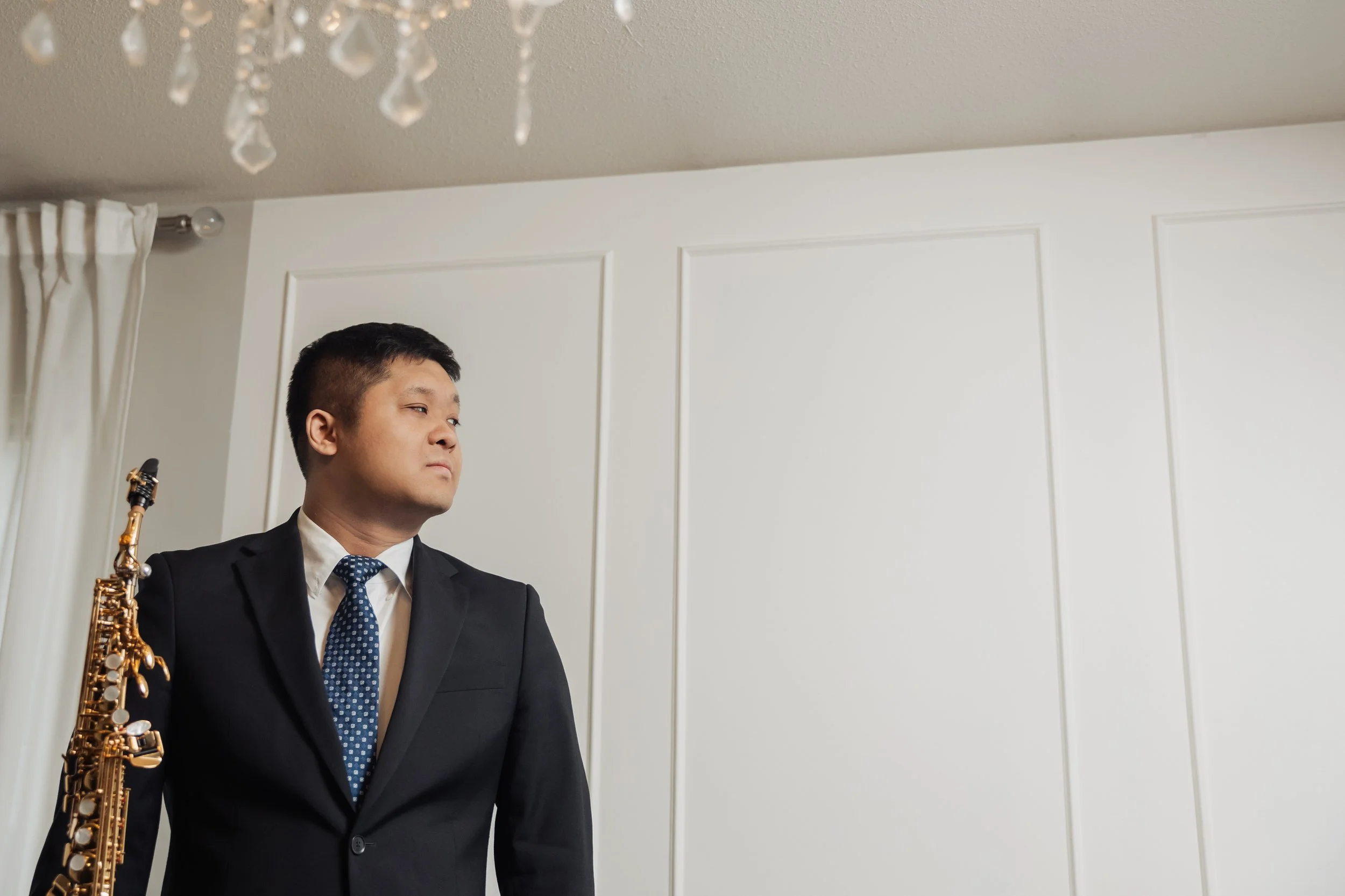 A man in a suit holding a saxophone, standing indoors near a wall with paneled white wainscoting and a ceiling with a crystal chandelier.