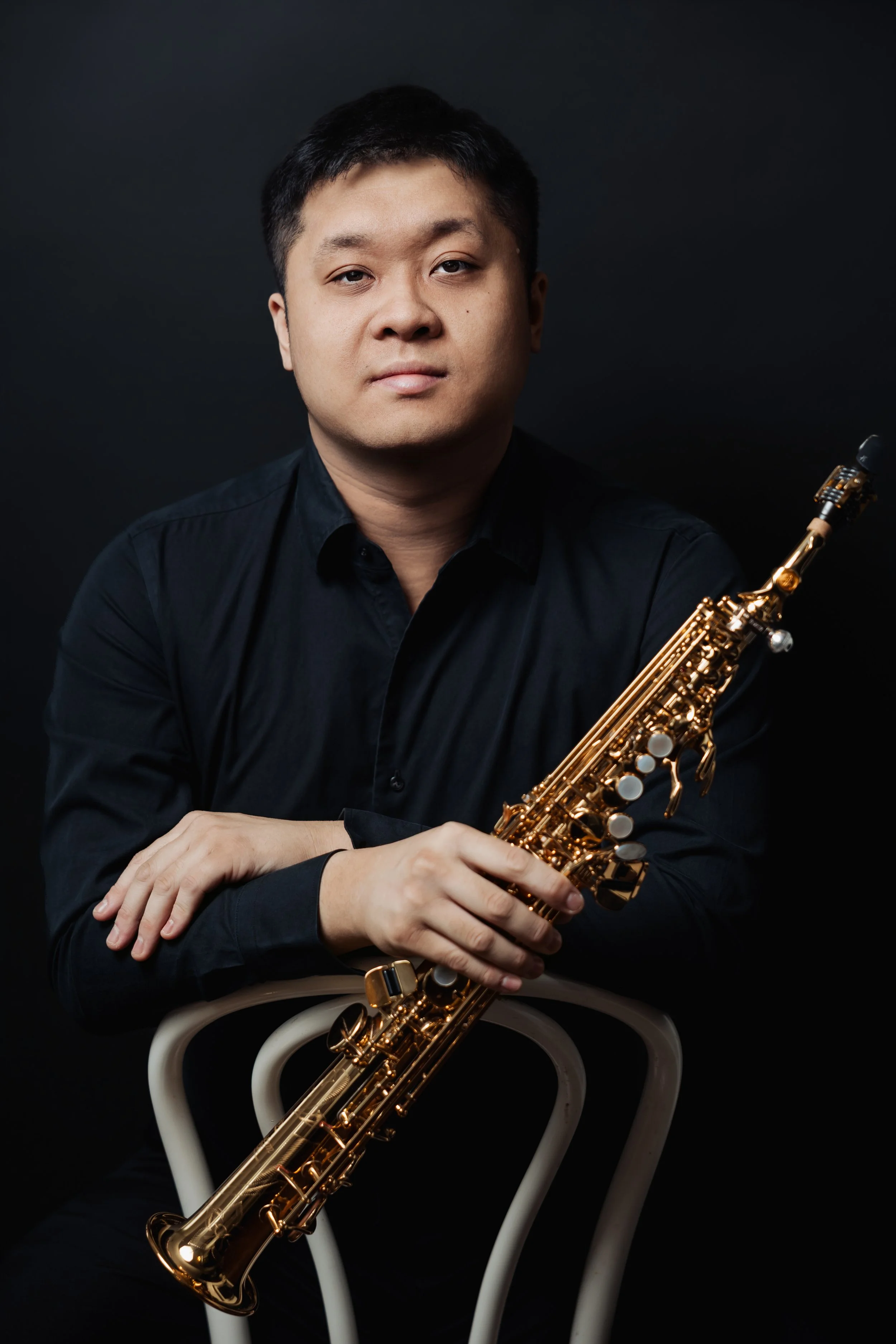 Portrait of a man in a black shirt holding a gold-colored saxophone, seated on a white chair against a dark background.
