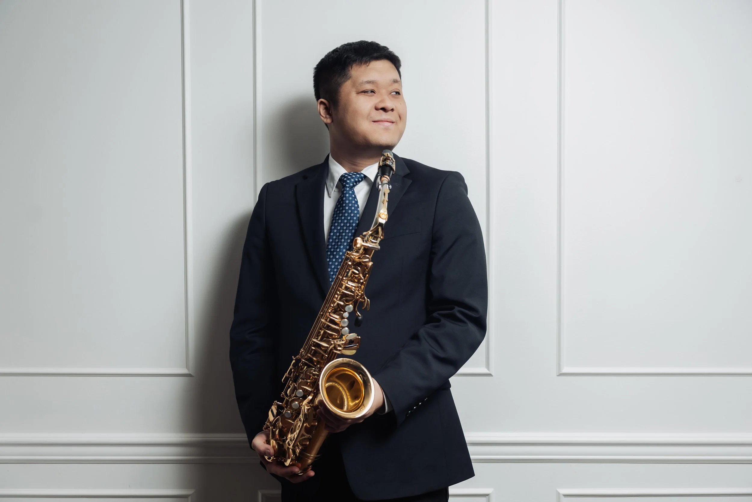 A man in a dark suit and blue patterned tie holding a gold-colored saxophone, standing against a white paneled wall, smiling slightly and looking to his left.