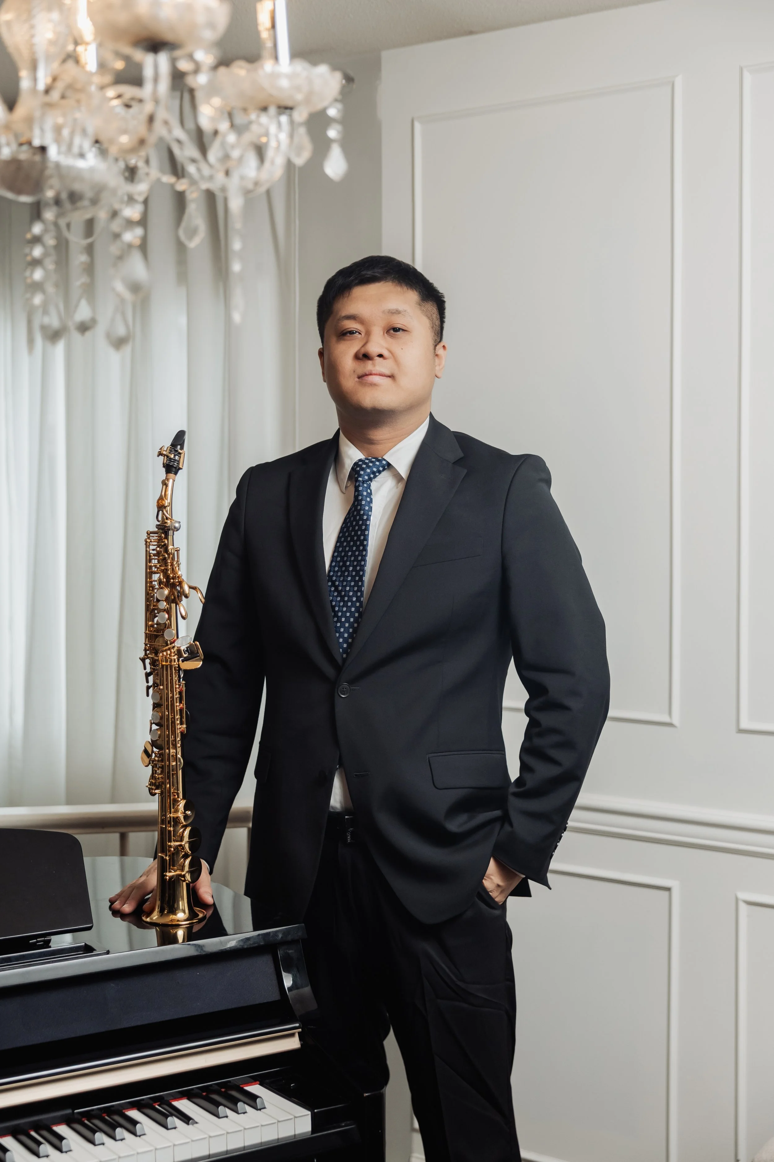 A man in a black suit and tie standing next to a piano and holding a gold saxophone in a room with white paneled walls and a chandelier.
