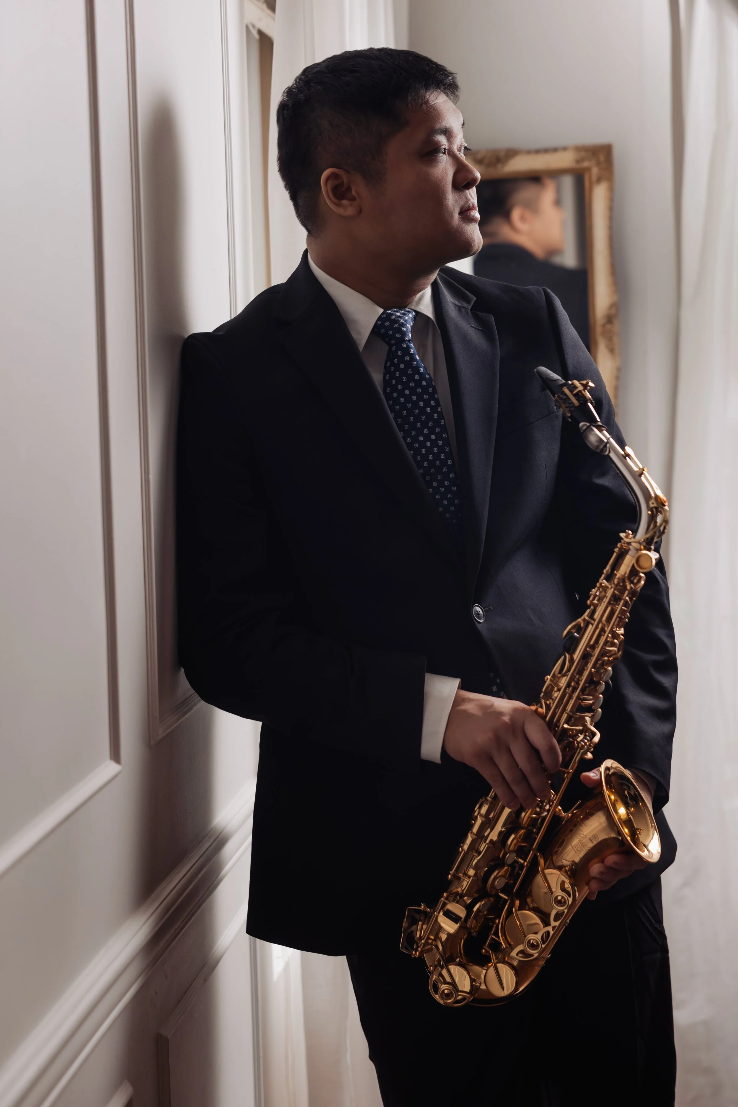 A man in a dark business suit holding a gold saxophone, standing in a white room with wall paneling, facing sideways, with a mirror in the background reflecting his face.