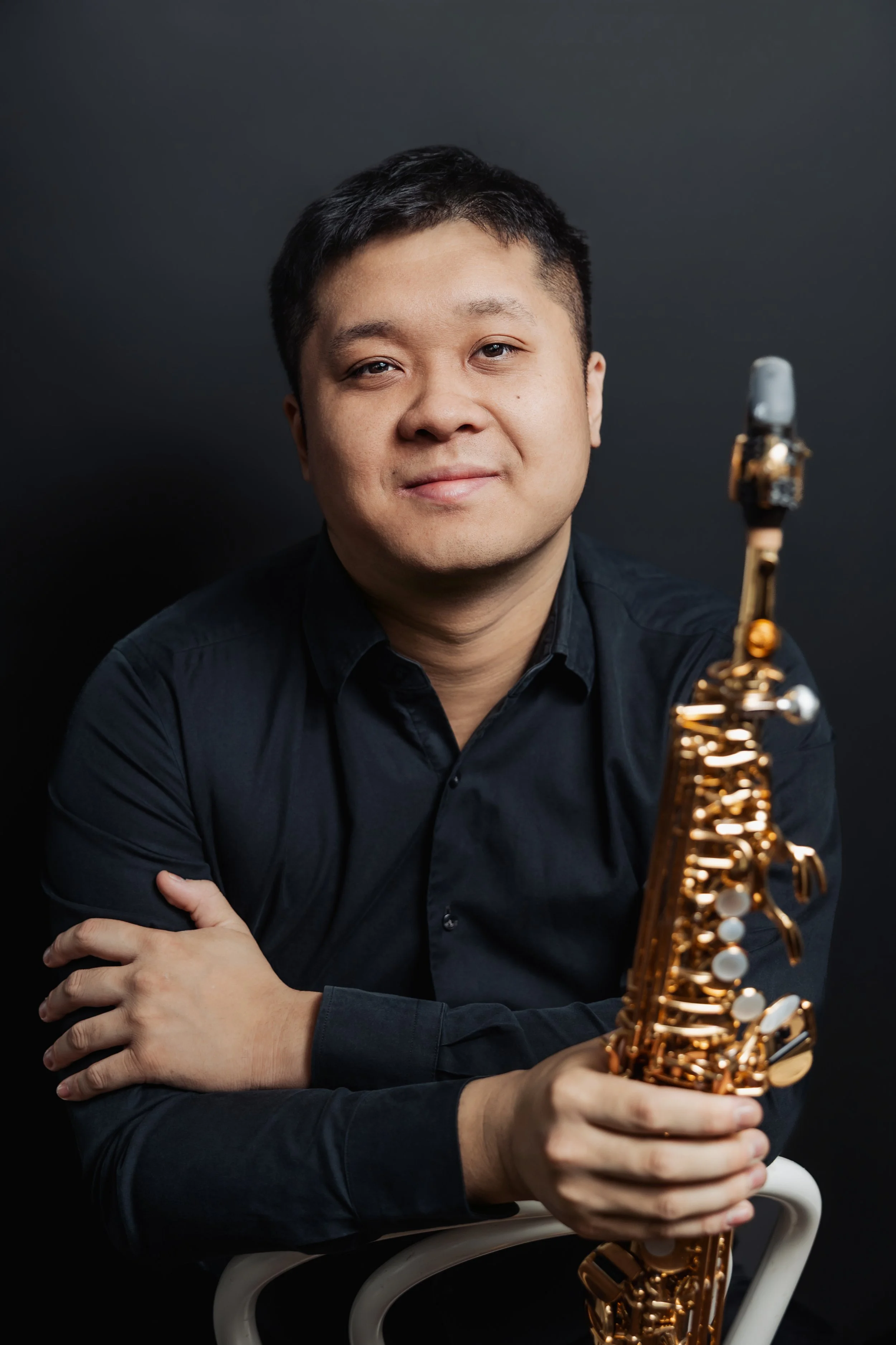 Portrait of a man with short black hair wearing a black shirt, sitting with arms crossed, holding a gold-colored saxophone, against a dark background.