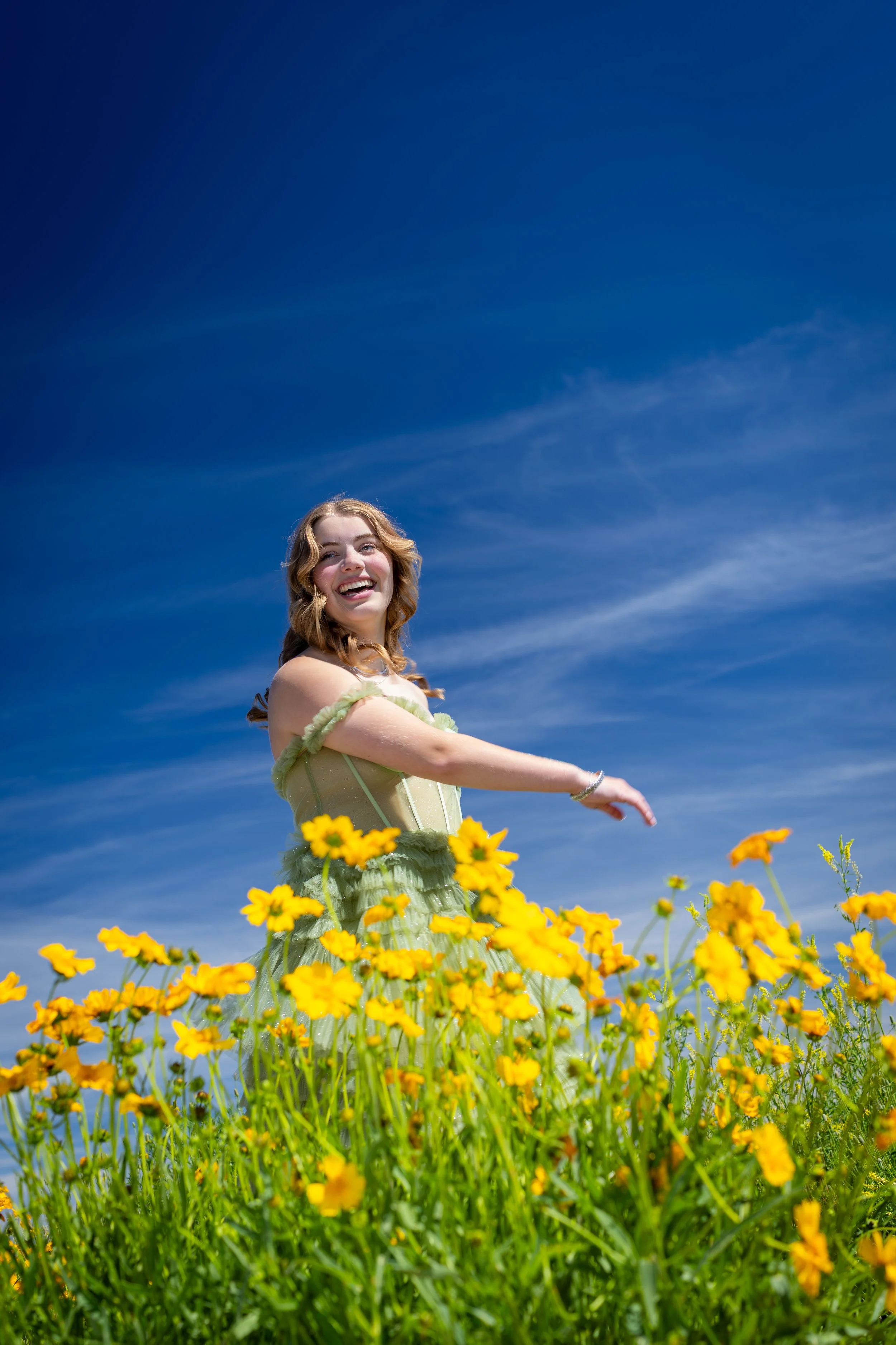 A woman smiling and wearing a light green dress, standing among yellow flowers in a bright outdoor setting with a blue sky and wispy clouds in Colorado. This is an example of a styled senior portait.