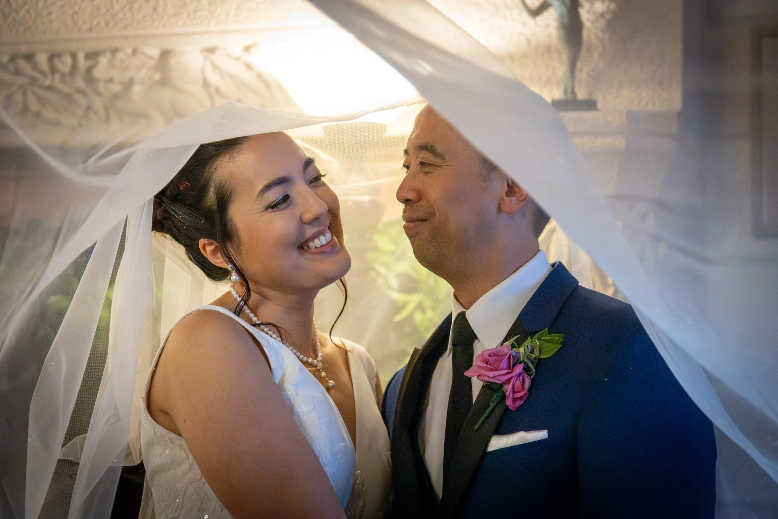 A bride and groom face each other under a wedding veil in a close-up shot. The bride has dark hair styled up, and is wearing pearl jewelry and a white dress. The groom is dressed in a dark suit with a pink boutonniere and a bowtie. They are smiling tenderly at each other, creating a romantic and joyful moment.