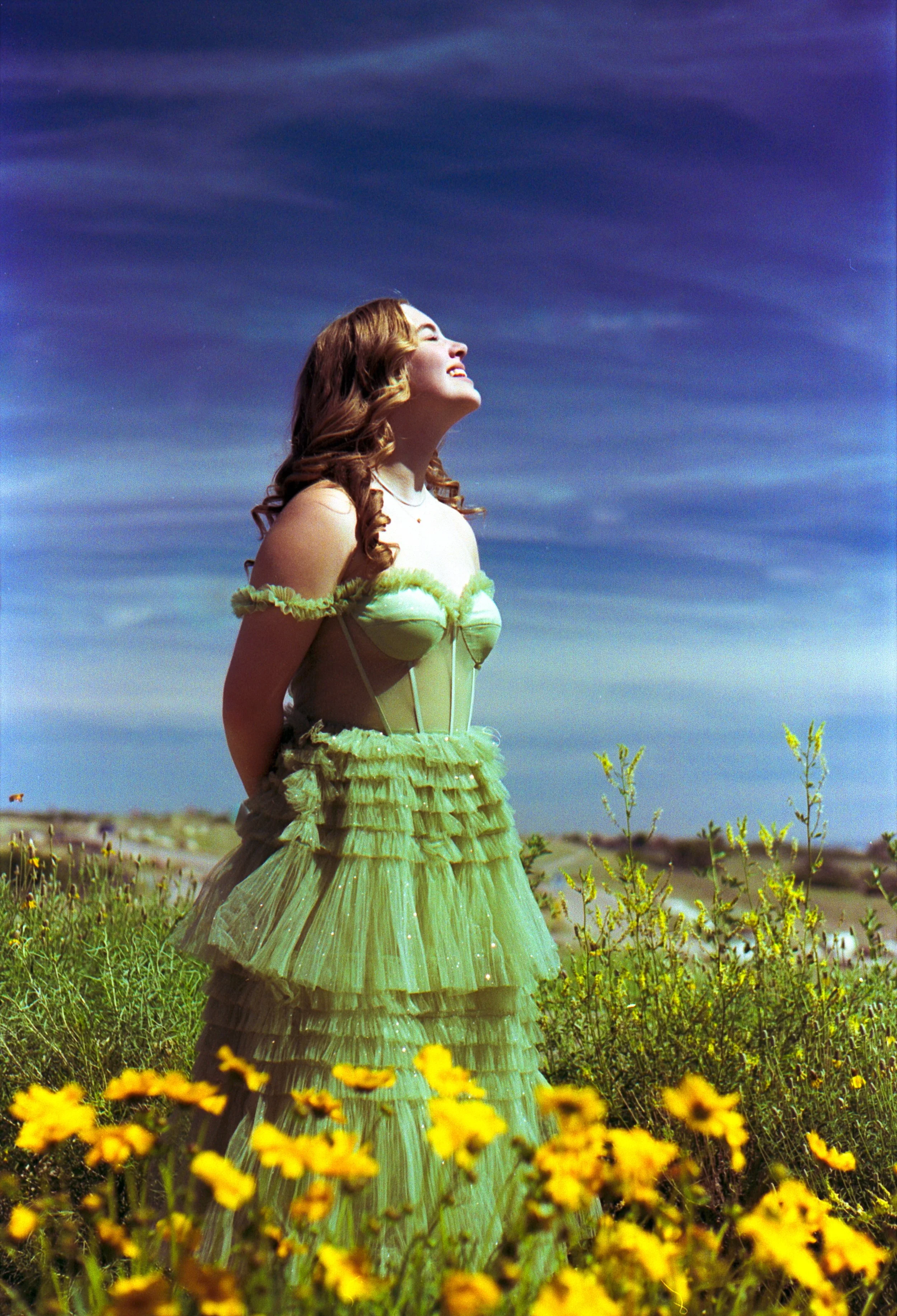 A high school senior with long, curly, hair is standing in a field of yellow flowers, smiling and looking up at the sky. She is wearing a light yellow, ruffled dress. This photo is an example of film photography in colorado