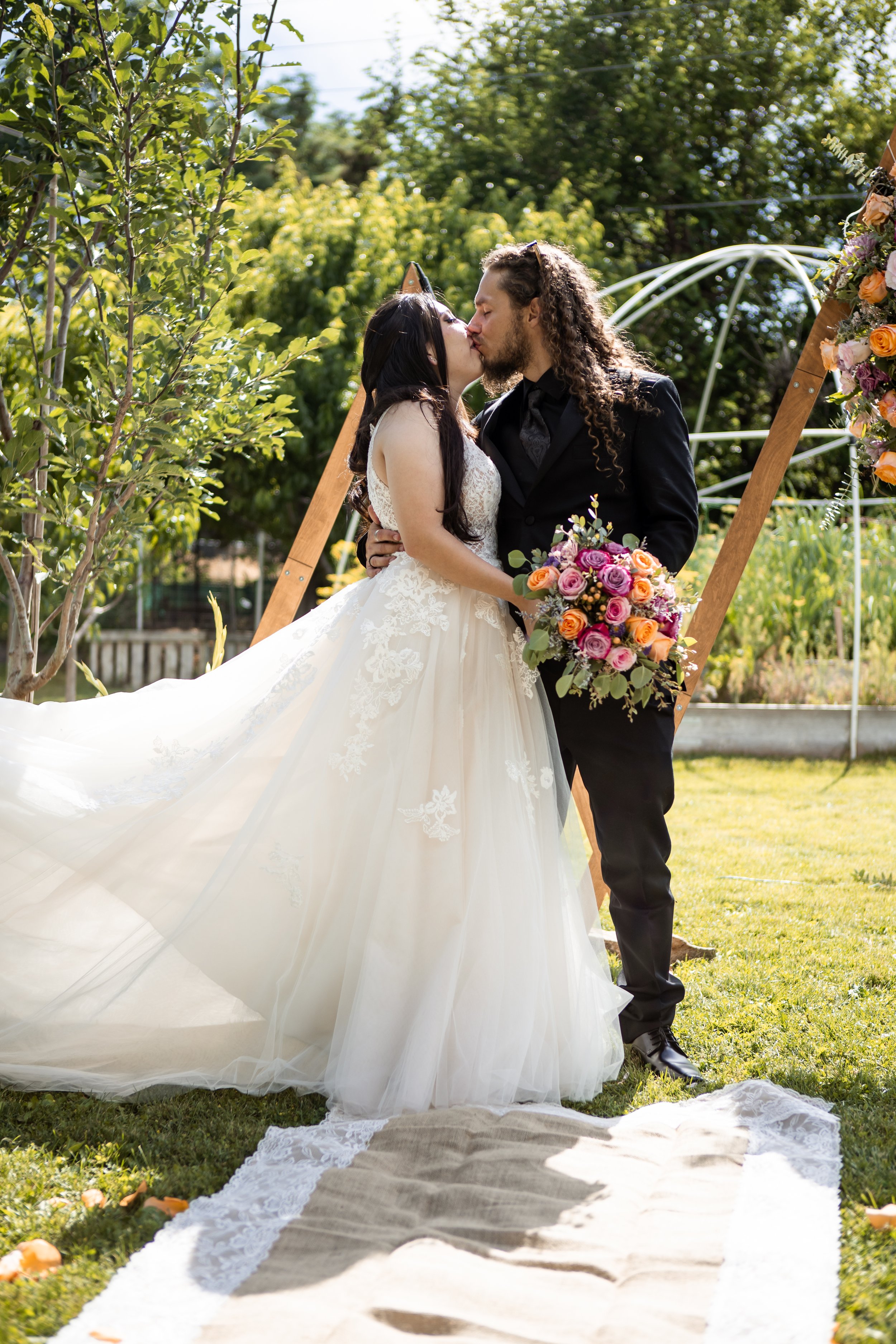 A bride and groom sharing a kiss outdoors in a colorado backyard wedding, with the bride holding a colorful bouquet of flowers, and a wedding arch decorated with flowers in the background.