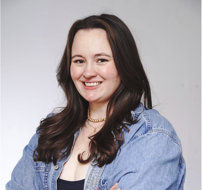 A young woman photographer smiling with long brown hair, wearing a denim jacket over a black top, and a gold beaded necklace.