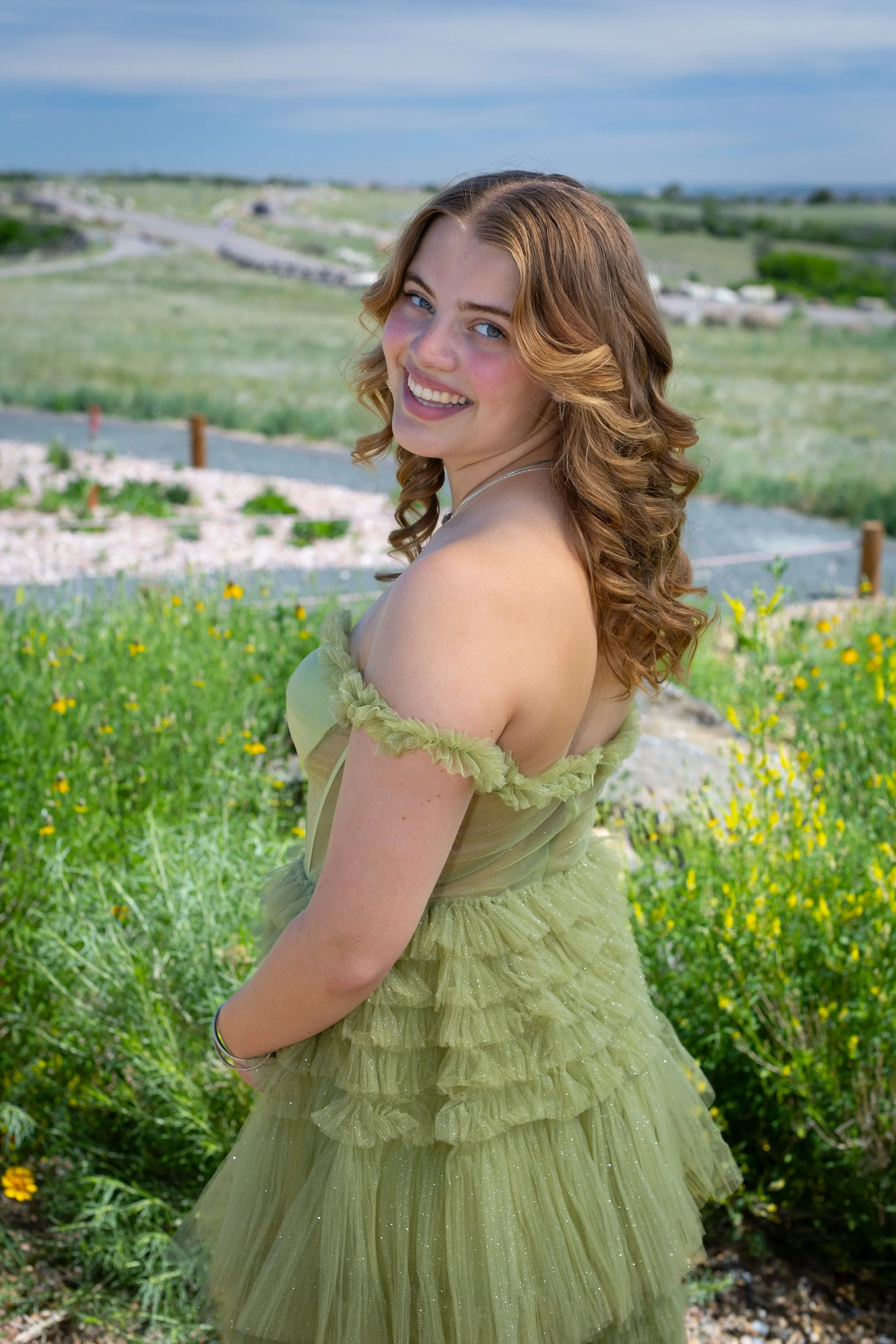 A senior with long, curly brown hair smiling outdoors, wearing a light green dress with layered ruffles, standing in a grassy area with yellow flowers, a gravel path, and a scenic landscape with rolling hills and a cloudy sky in the background.