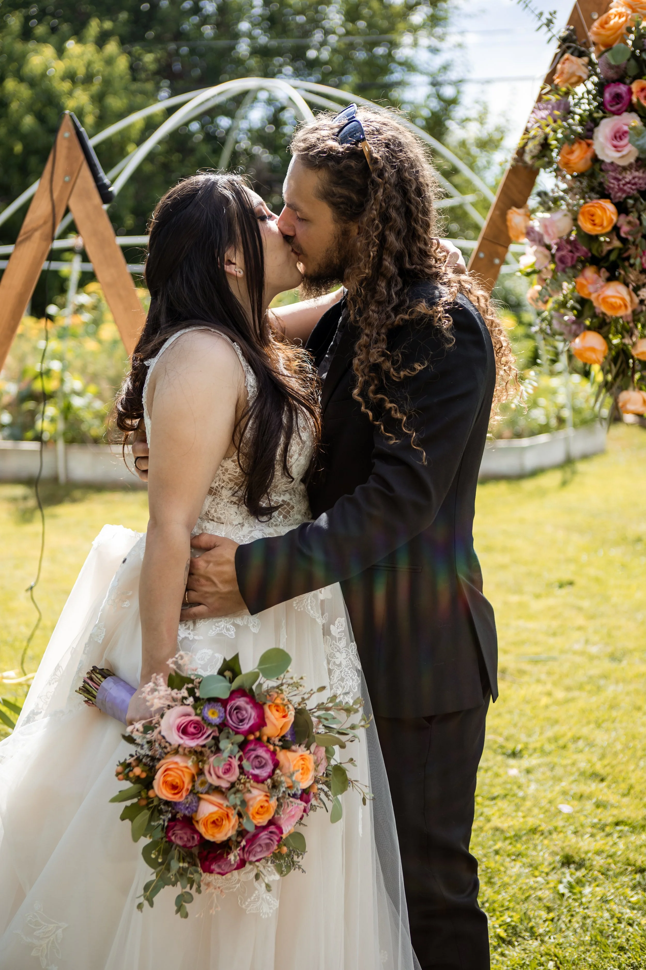 A bride and groom kiss during their outdoor colorado wedding ceremony, with trees and a floral arch in the background. The bride holds a bouquet of pink, orange, and purple flowers, while the groom wears a black suit.