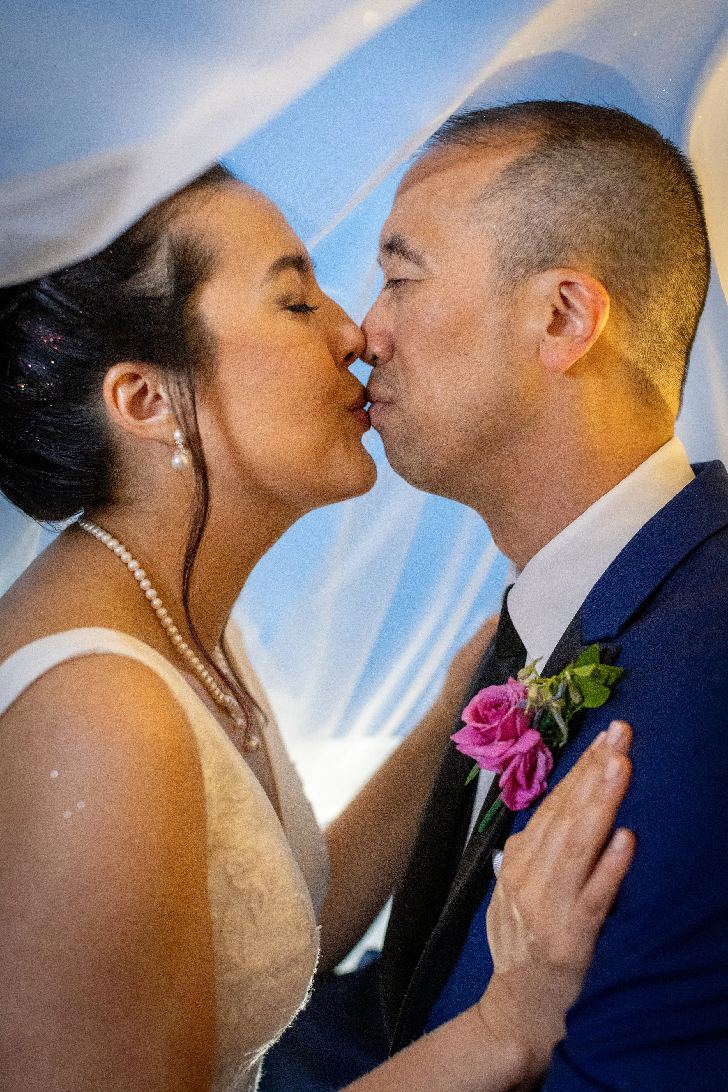 An asian bride and groom kiss under the brides veil illuminated with warm glow. Sparkles in the brides hair catch the light. The portrait captures the perfect moment of wedding day bliss.