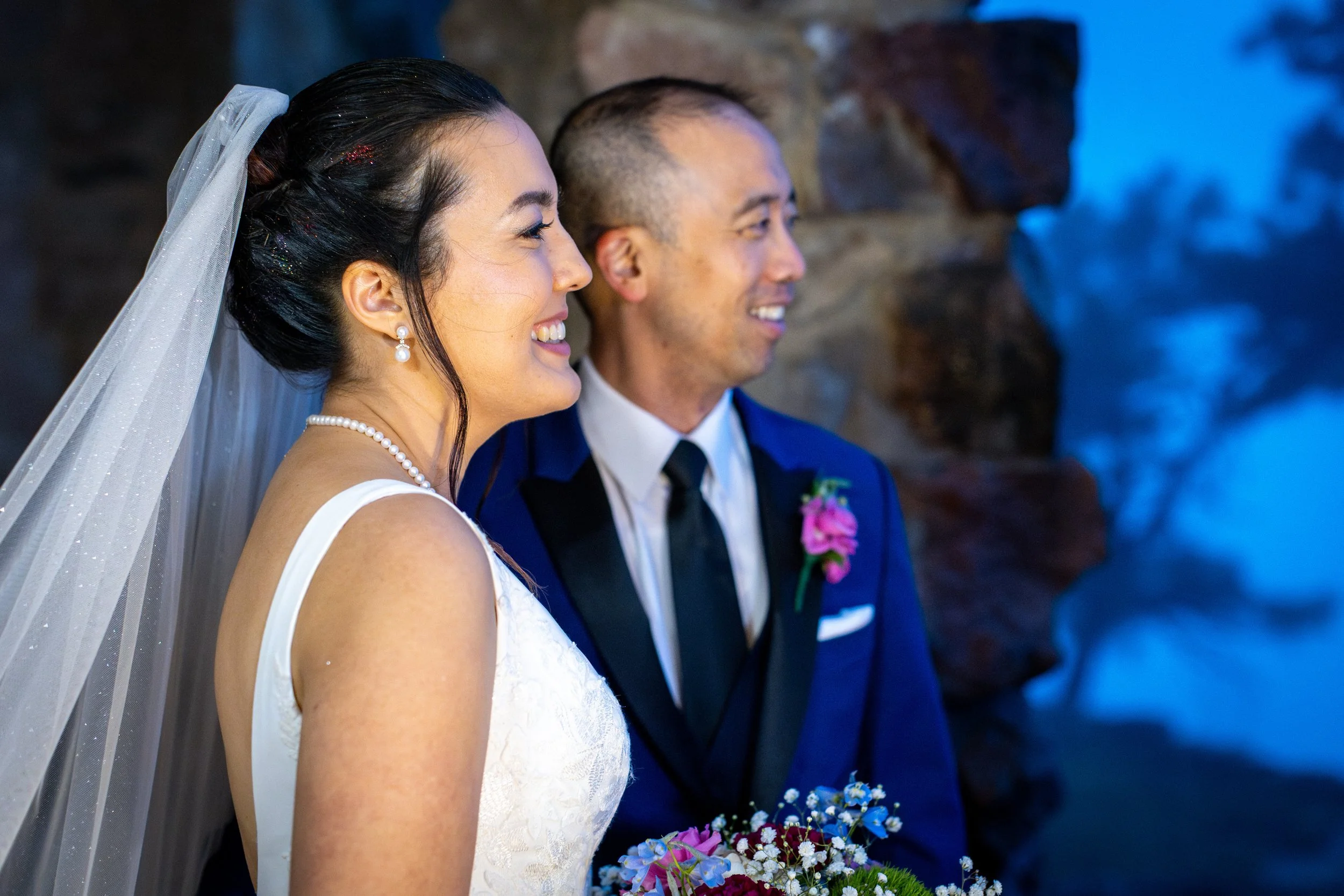 A bride with dark hair and pearl earrings smiling, standing next to a groom in a blue suit with a boutonniere, holding a bouquet of colorful flowers, at a wedding ceremony in Colorado.