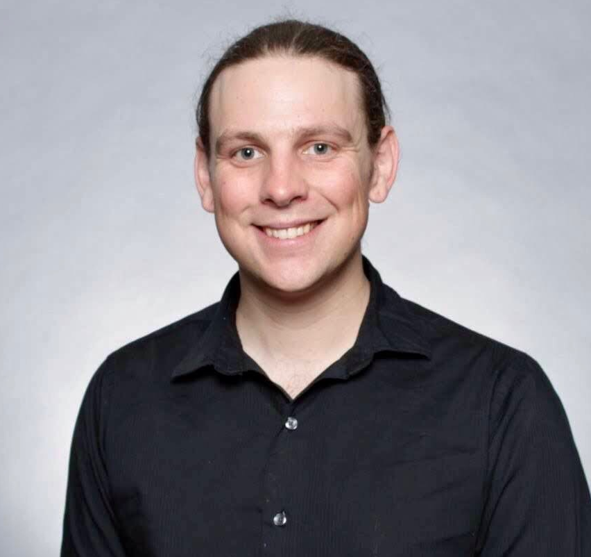 A young man videographer with light skin, brown hair, and blue eyes, smiling, wearing a black button-up shirt against a plain light gray background.