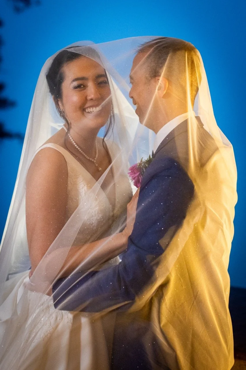 A bride and groom are smiling and close together under a wedding veil during their wedding ceremony in Colorado.