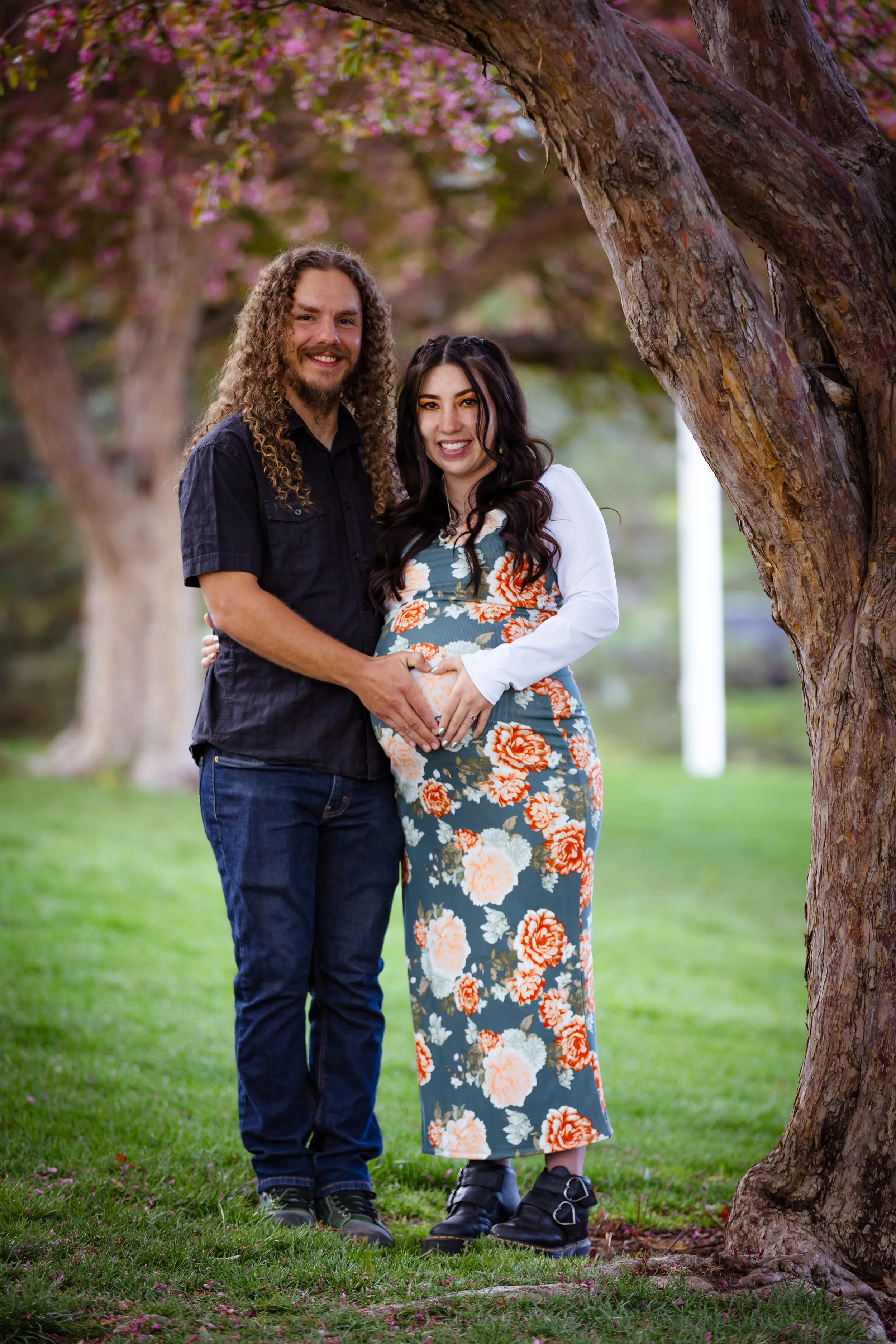 A happy couple standing outdoors under a tree, with the woman appearing pregnant, smiling and holding her belly, in a park with pink blooming trees. This is a colorado maternity photoshoot.