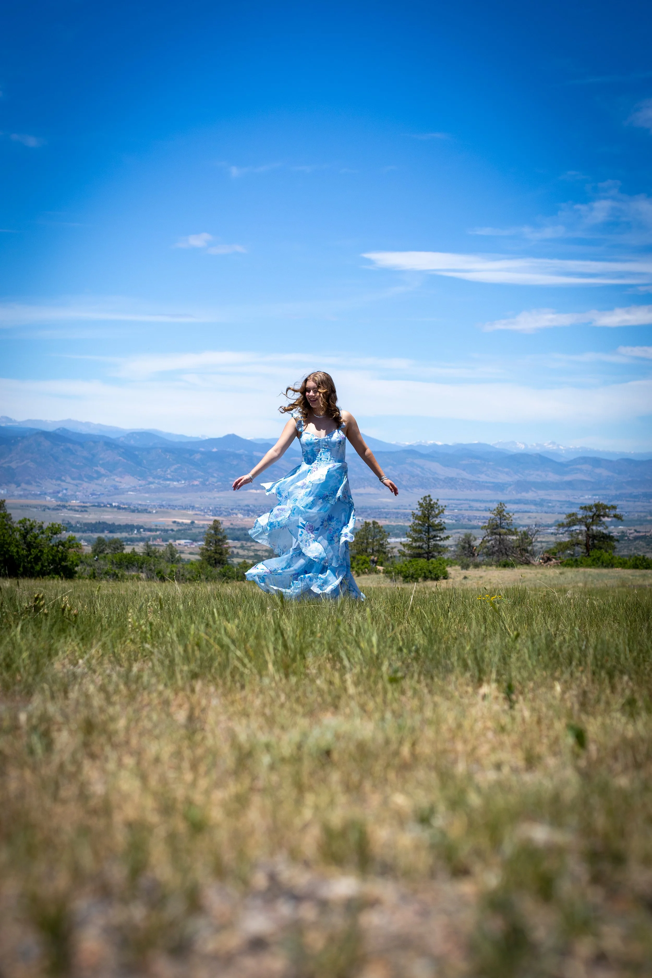A woman in a flowing blue dress is joyfully jumping in a grassy field with mountains in the background and a partly cloudy sky overhead. This is an example of Colorado senior photos with rolling mountain vistas.