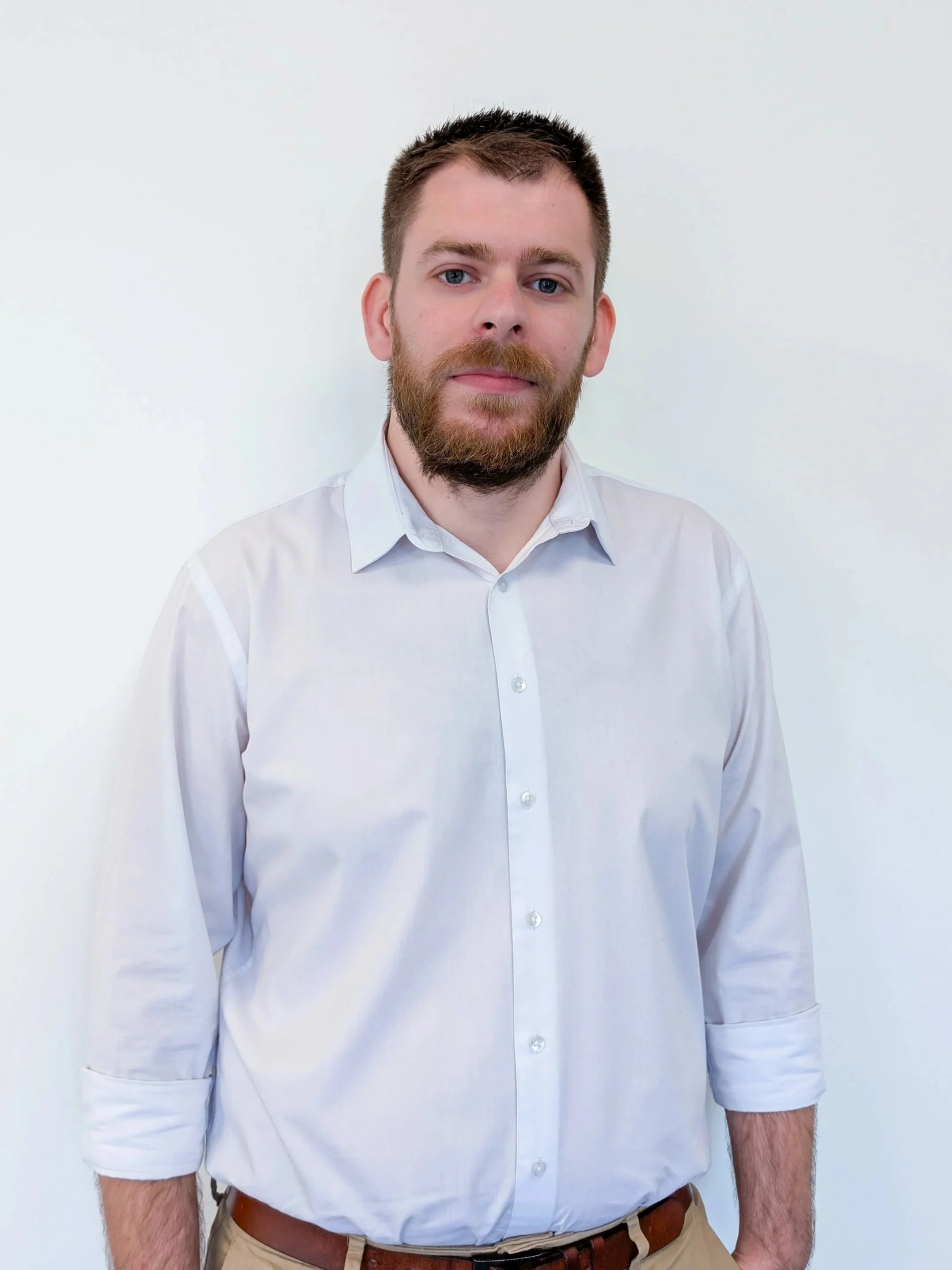 A man with a beard and short brown hair wearing a white button-up shirt with rolled-up sleeves and beige pants standing against a plain light-colored background.