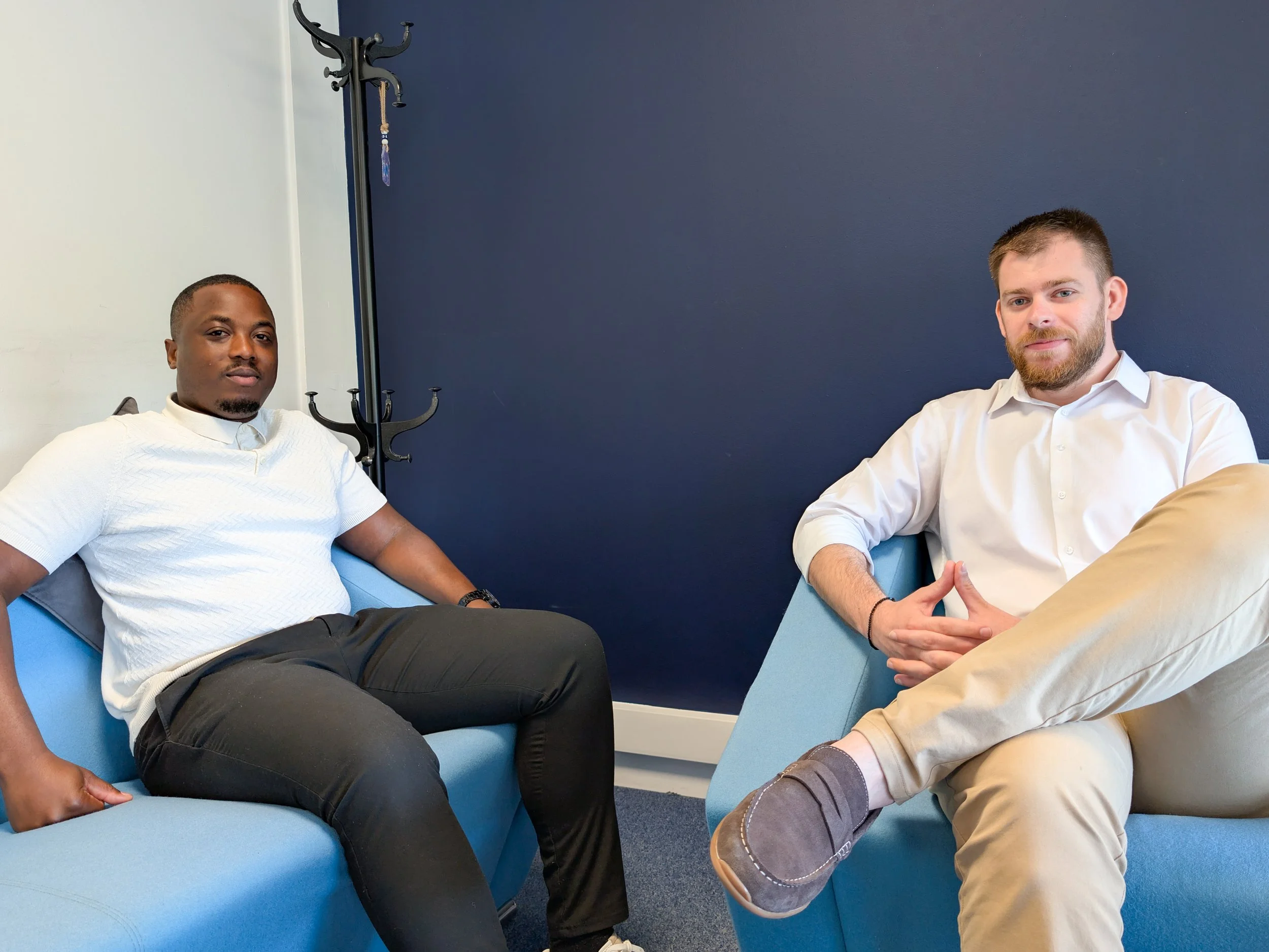 Two men sitting on blue couches in a room with a dark blue wall. One man on the left wears a white shirt and black pants, the man on the right wears a white shirt and beige pants. They are holding hands with interlocked fingers.