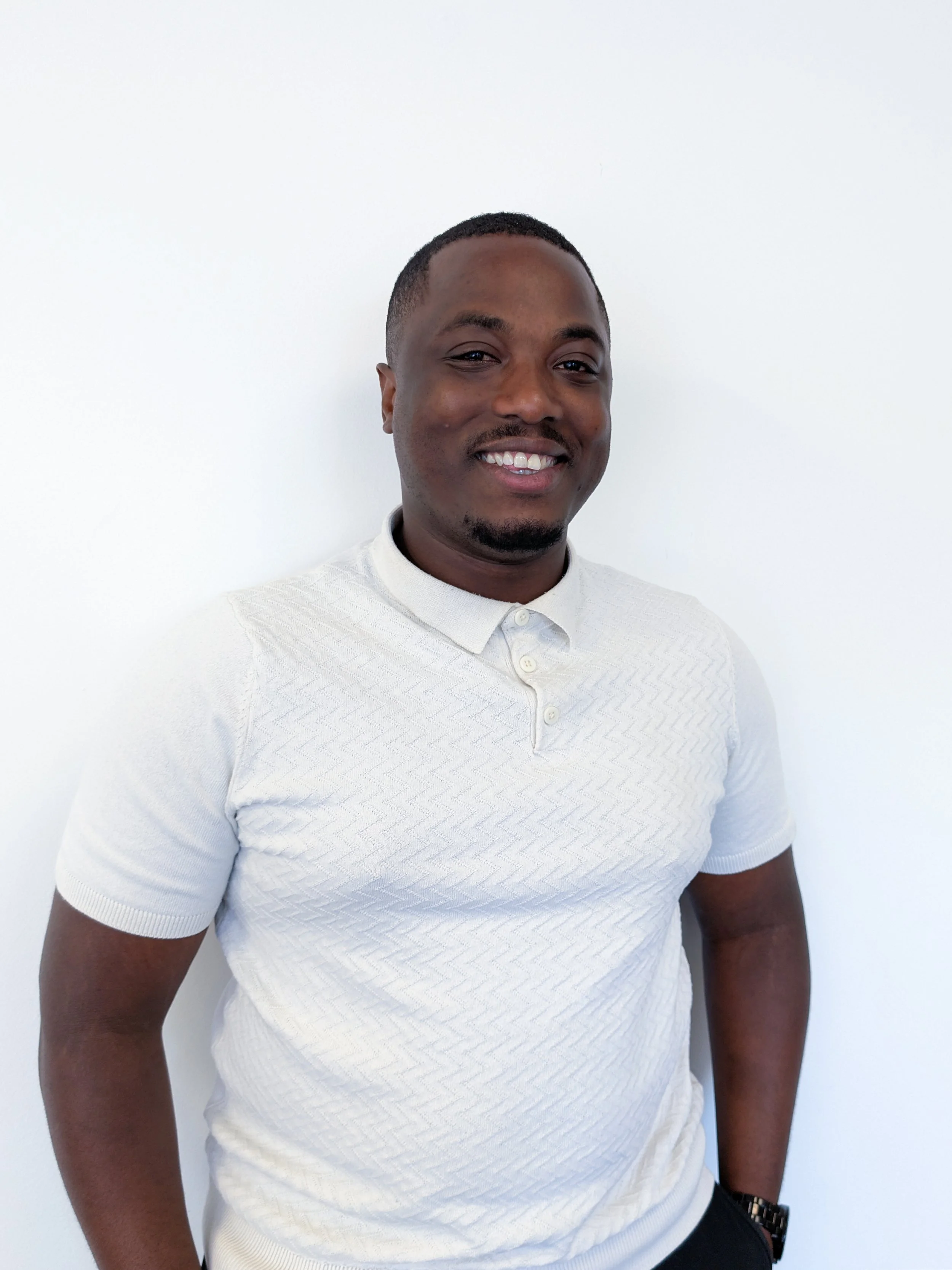 A man with short hair and a goatee smiling, wearing a white textured polo shirt and a watch, standing against a plain white wall.