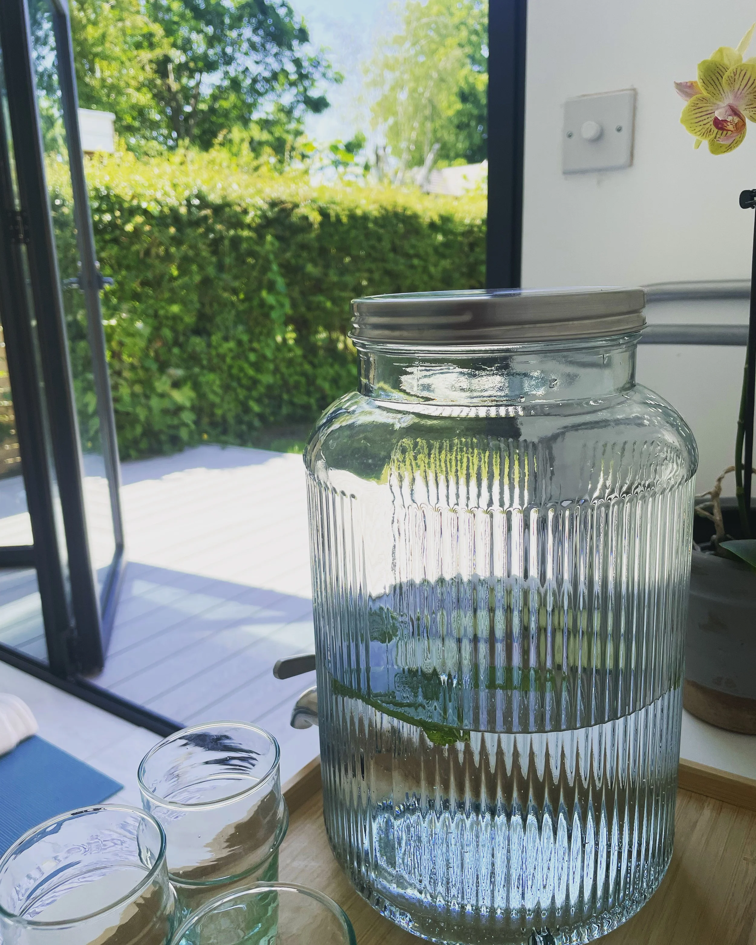 A clear glass jar with a metal lid on a wooden surface, with smaller glass containers in front, placed near a window showing a bright outdoor scene with green trees and a blue sky.