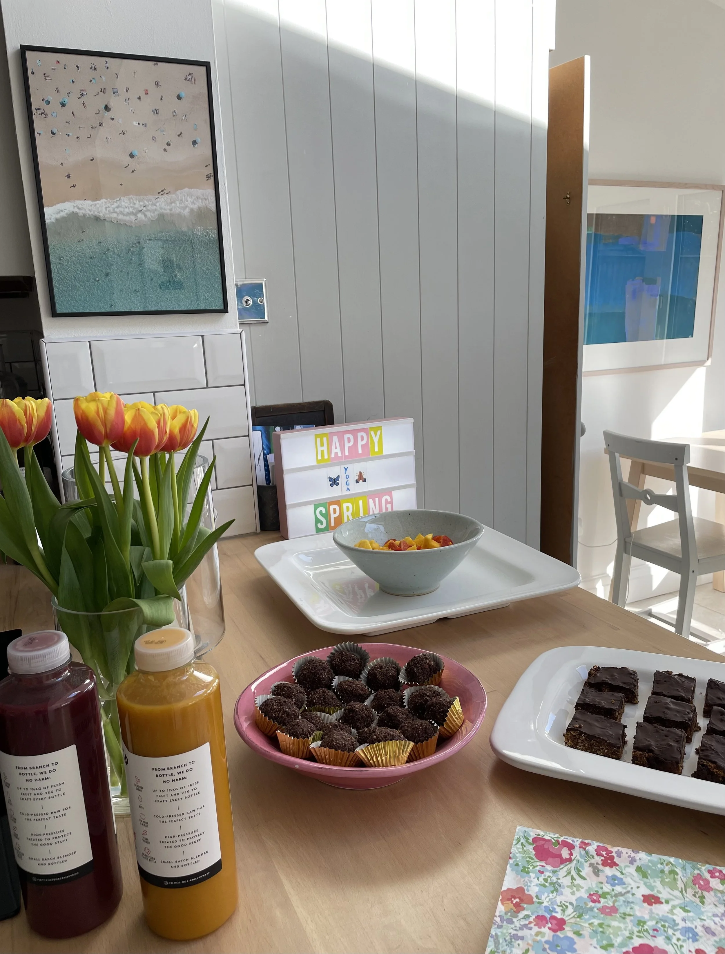 A table decorated for spring featuring a bouquet of yellow and pink tulips, bottles of juice, and various desserts including chocolate cake squares, chocolate truffles, and fruit salad. There are also decorative signs with the words "HAPPY SPRING." In the background, wall art displays a beach scene with umbrellas and people on the sand.