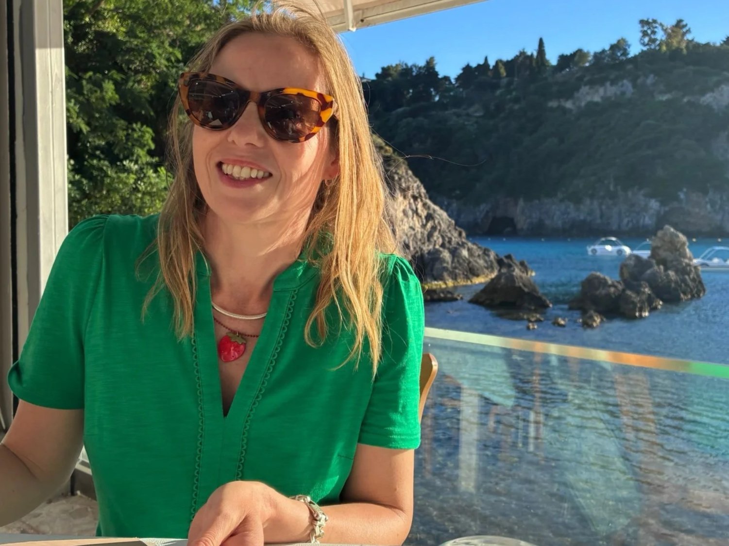 A woman with blonde hair wearing sunglasses and a green shirt, sitting in front of a scenic coastal background with rocks and water, smiling.