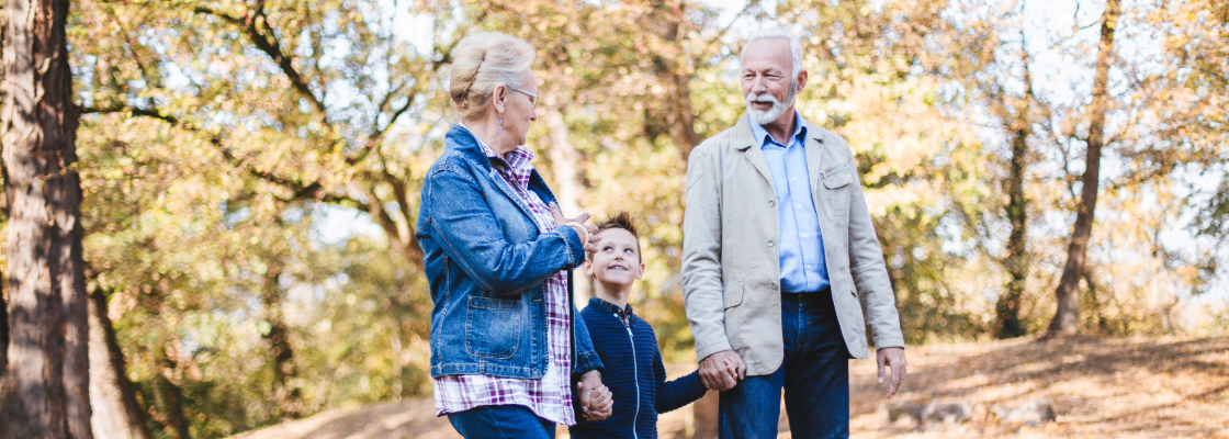 Grandparents and a child walking outdoors in a park with autumn trees.
