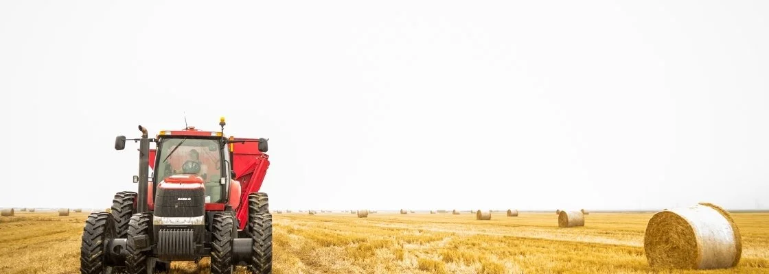 A red tractor in a field with large hay bales scattered across the golden harvest landscape under a cloudy sky.