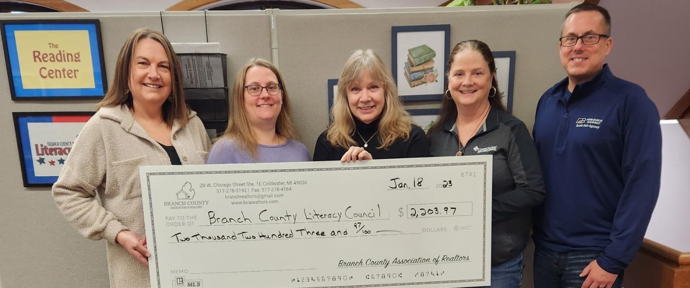 Five people standing indoors, holding a large check from the Branch County Association of Realtors made out to Branch County Literacy Council for $2,203.97. There are framed pictures on the wall behind them, and a sign that says "The Reading Center."