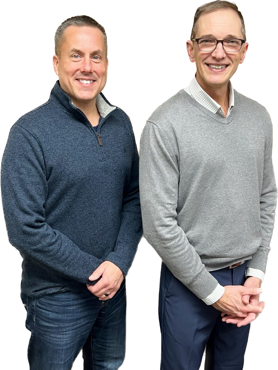 Two smiling men standing next to each other, one wearing a dark blue sweater and jeans, the other in a gray sweater and blue pants, with a plain background.