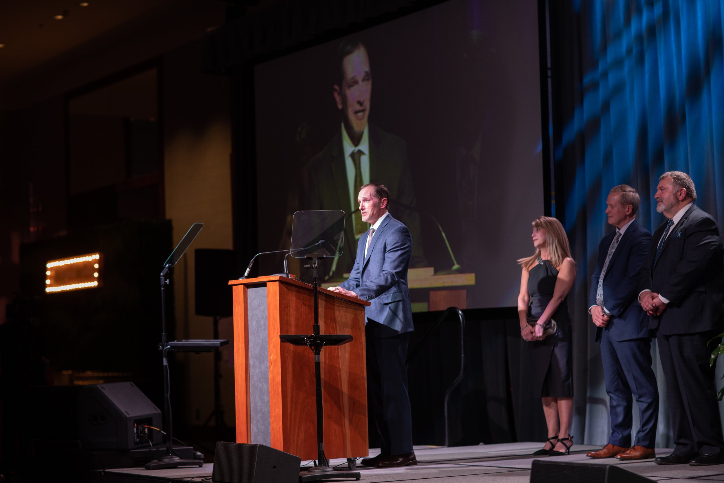 A man in a blue suit giving a speech at a podium with four people standing in a line behind him on stage. There is a large screen behind the speaker projecting his image.