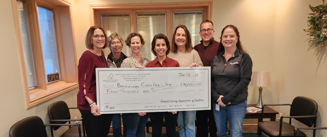 Group of eight women and one man holding a large ceremonial check in a room with beige walls and windows, celebrating a donation of $4,000 to Think Big Foundations.