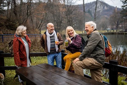 Four people, three women and one man, are outdoors by a lake on a cloudy day. They are smiling and holding drinks, engaging in conversation and laughter, with trees and mountains in the background.