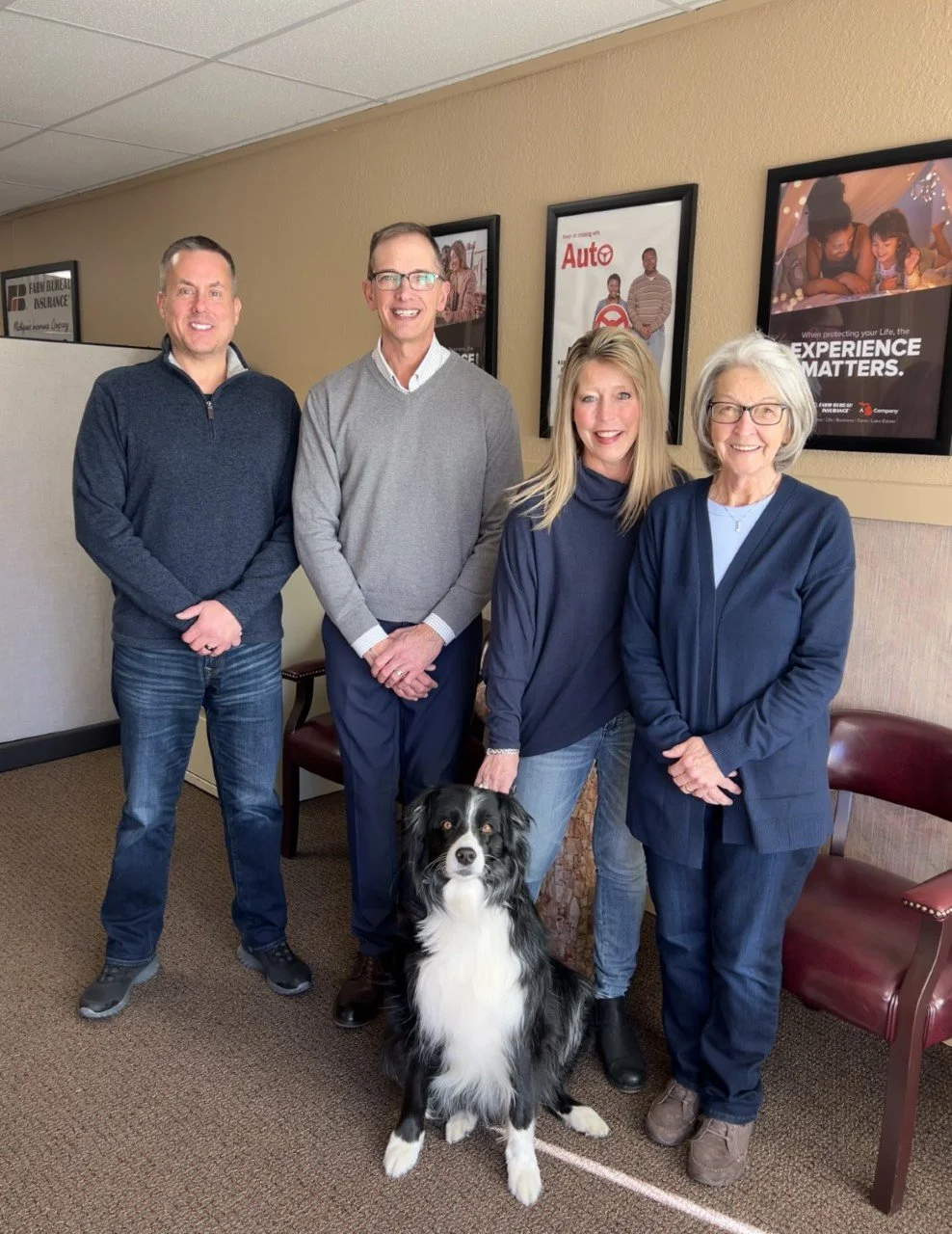 Four people standing with a black and white dog in an office waiting area. Three women and one man, all smiling, are dressed casually. Behind them are three framed posters on the wall. There are two chairs visible on either side of the group.